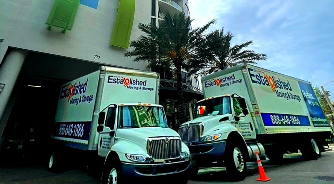 two established moving & storage trucks wait outside a high-rise