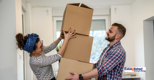 A couple walking into their new home. The man is holding a stack of boxes while the woman helps him balance said boxes.